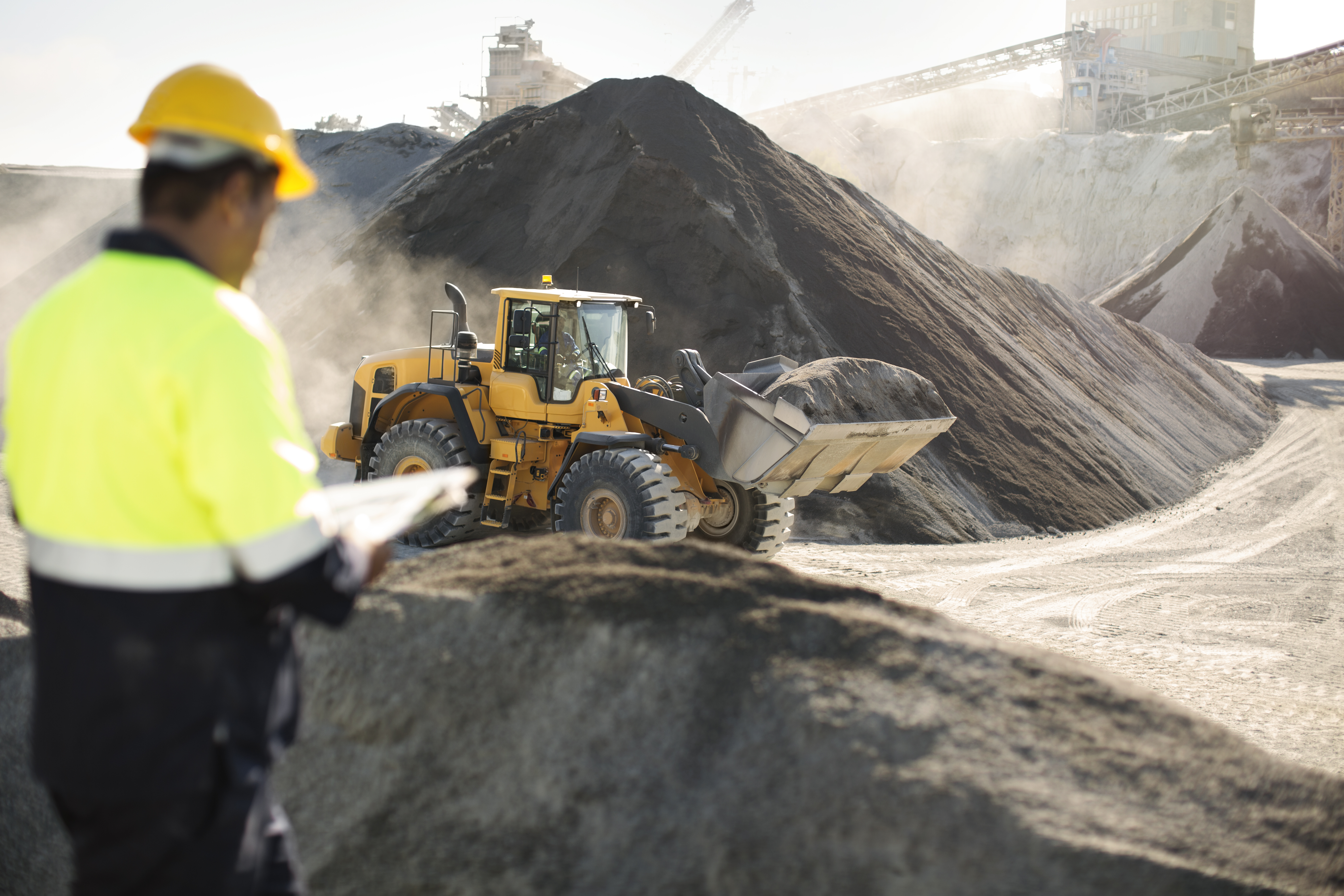 Quarry worker overseeing a loader operating near large mineral stockpiles at a mining site. 